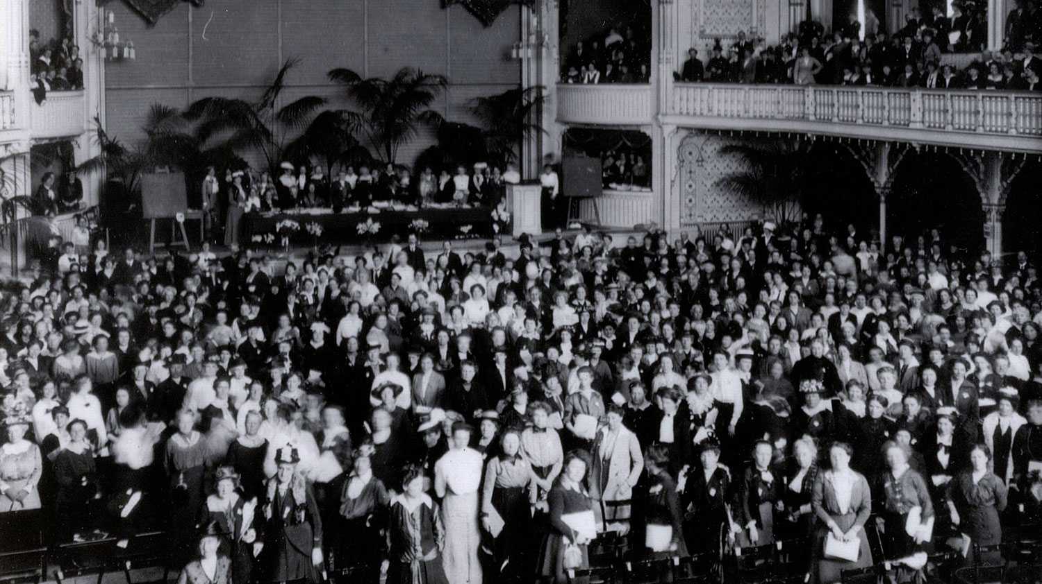 Many women photographed in huge auditorium facing stage.